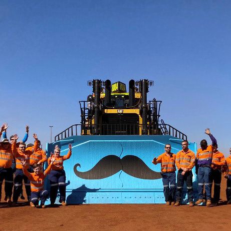 Photo of personnel at a mining site, leaping jubilantly in front of a tractor bearing a stylised Movember moustache logo.