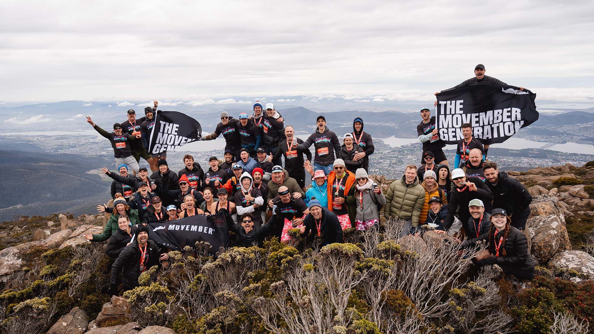 Photo of cheering people, holding a Movember flag, celebrating the completion of a half-marathon run at the summit of kunanyi/Mount Wellington in Hobart.