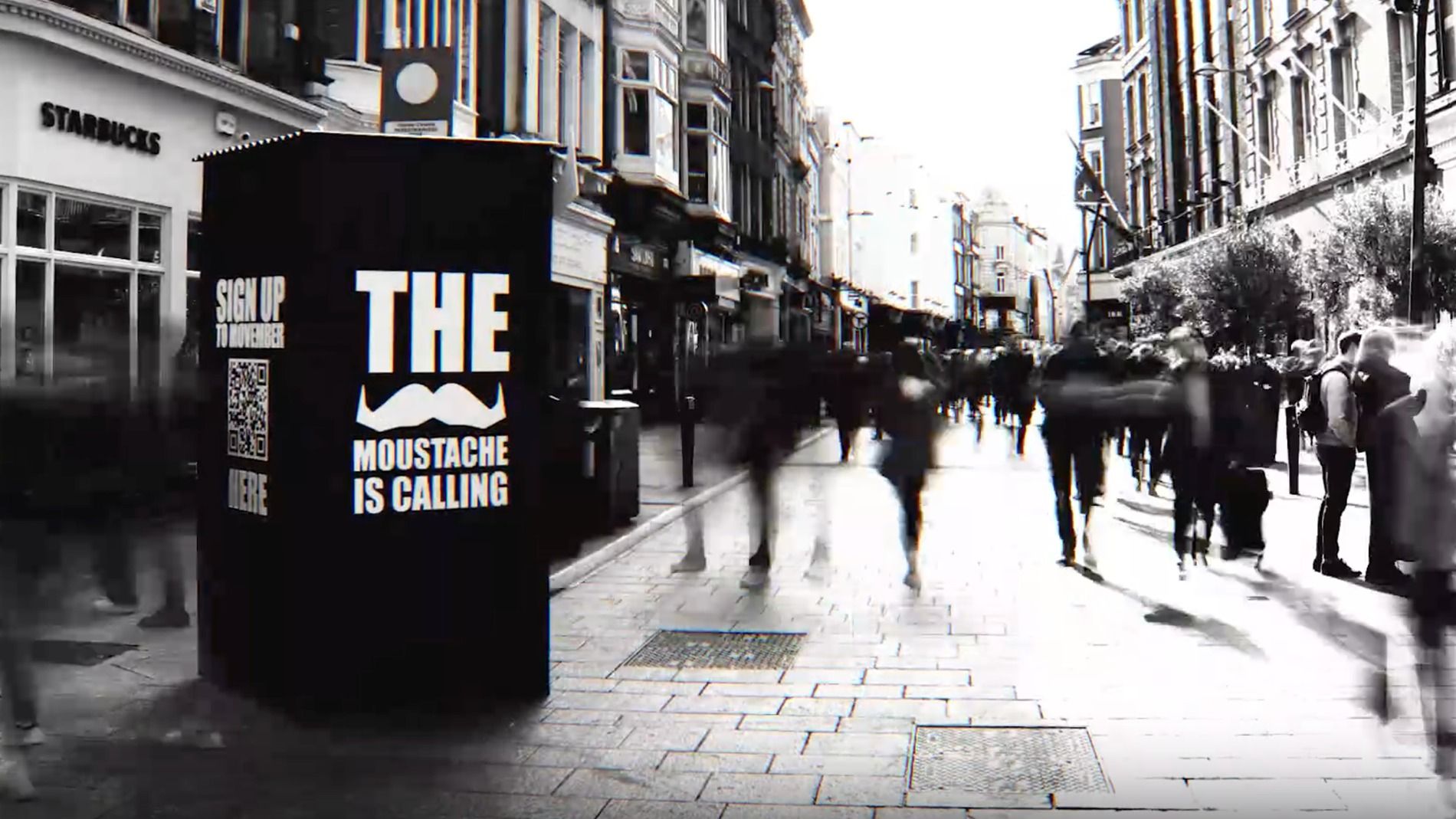 Black and white photo of a phone booth on a public street. Text on the phone booth reads: "The moustache is calling".