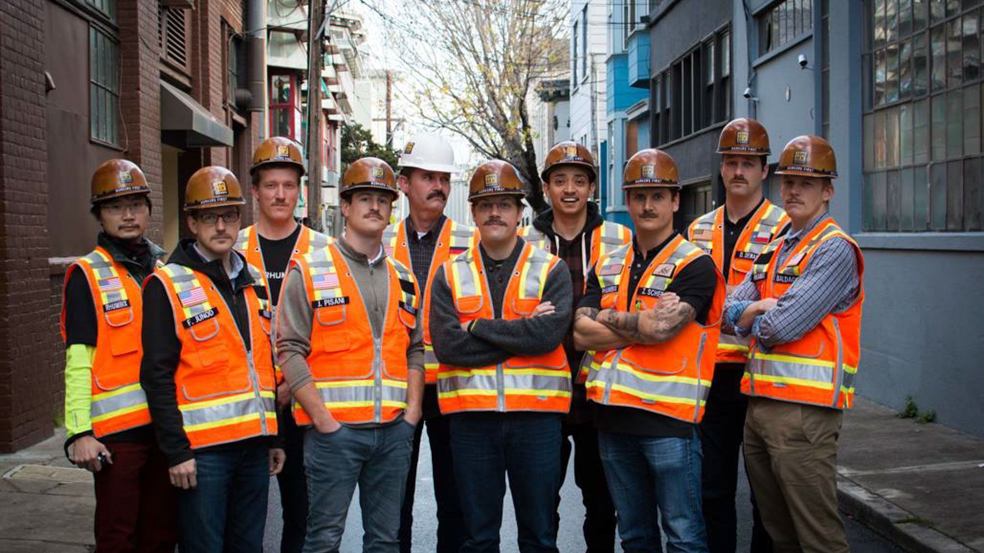 Group of men with high vis jackets and hard hats pose down alley way looking at camera 