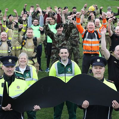 Photo of a group of emergency personnel in uniform, cheering to camera. Several participants in the foreground carry a large, black, moustache-shaped sign.