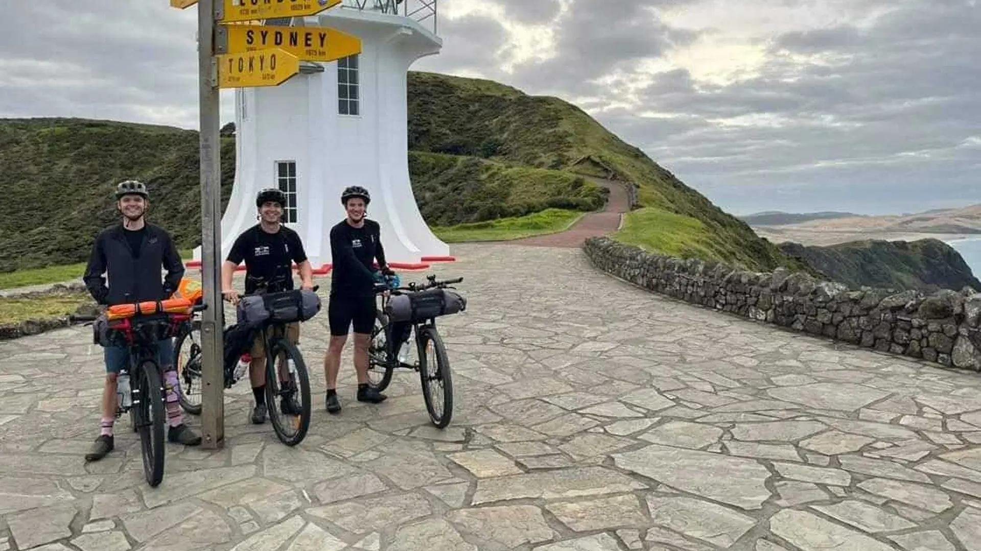 Three friends in riding attire, posing with their bicycles before a lighthouse.