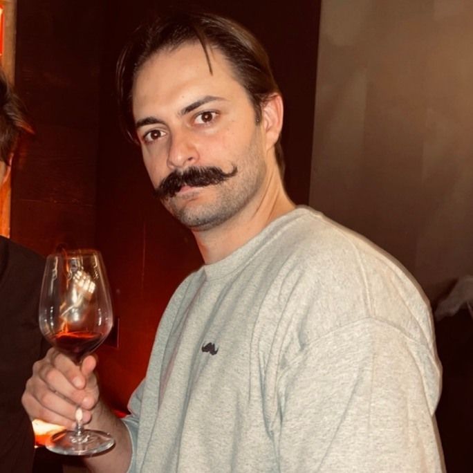 Young man sporting an excellent twirled moustache, holding a glass to camera.