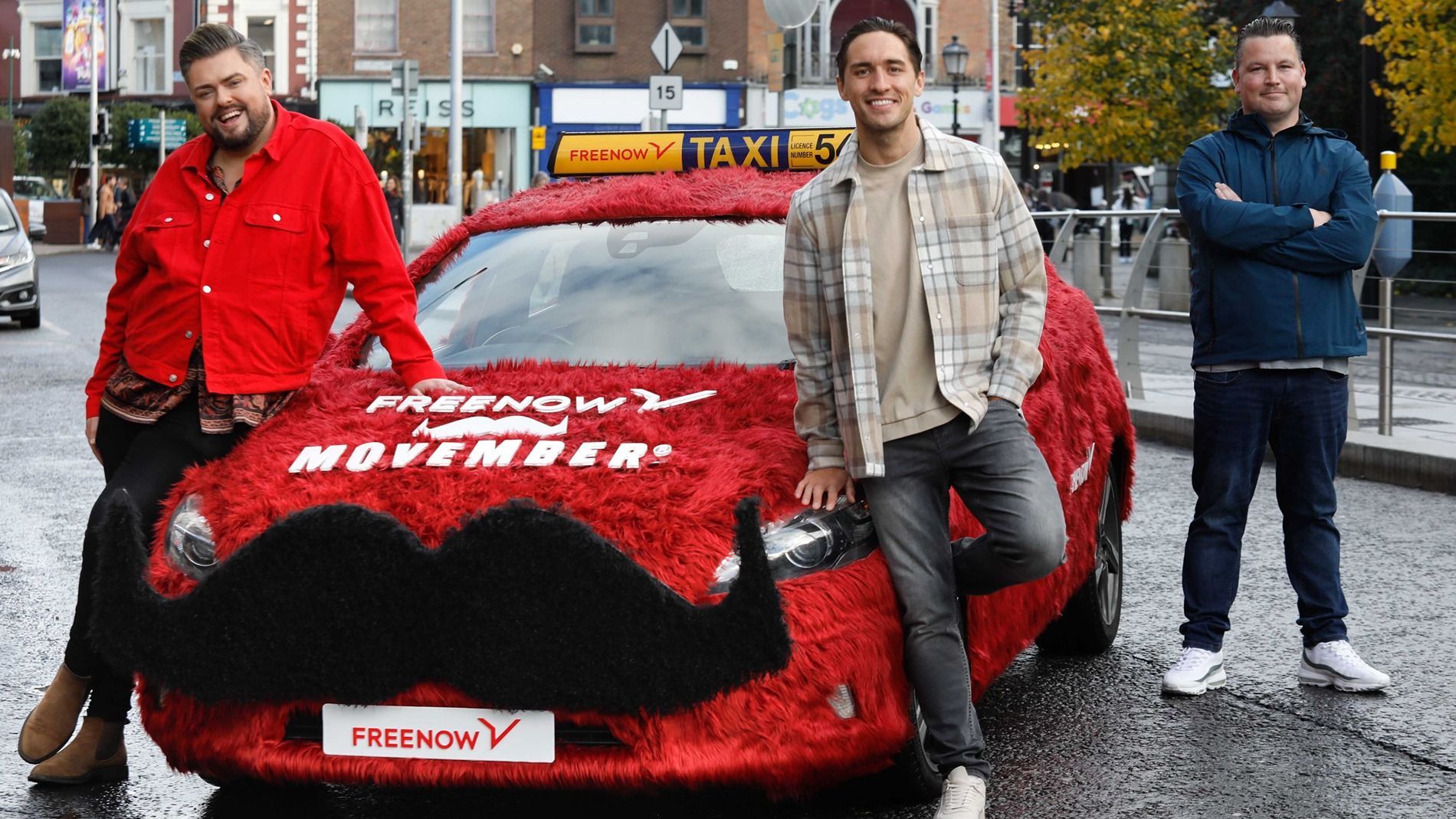 3 men stood outside a red hairy FREENOW taxi with a black Movember moustache on it