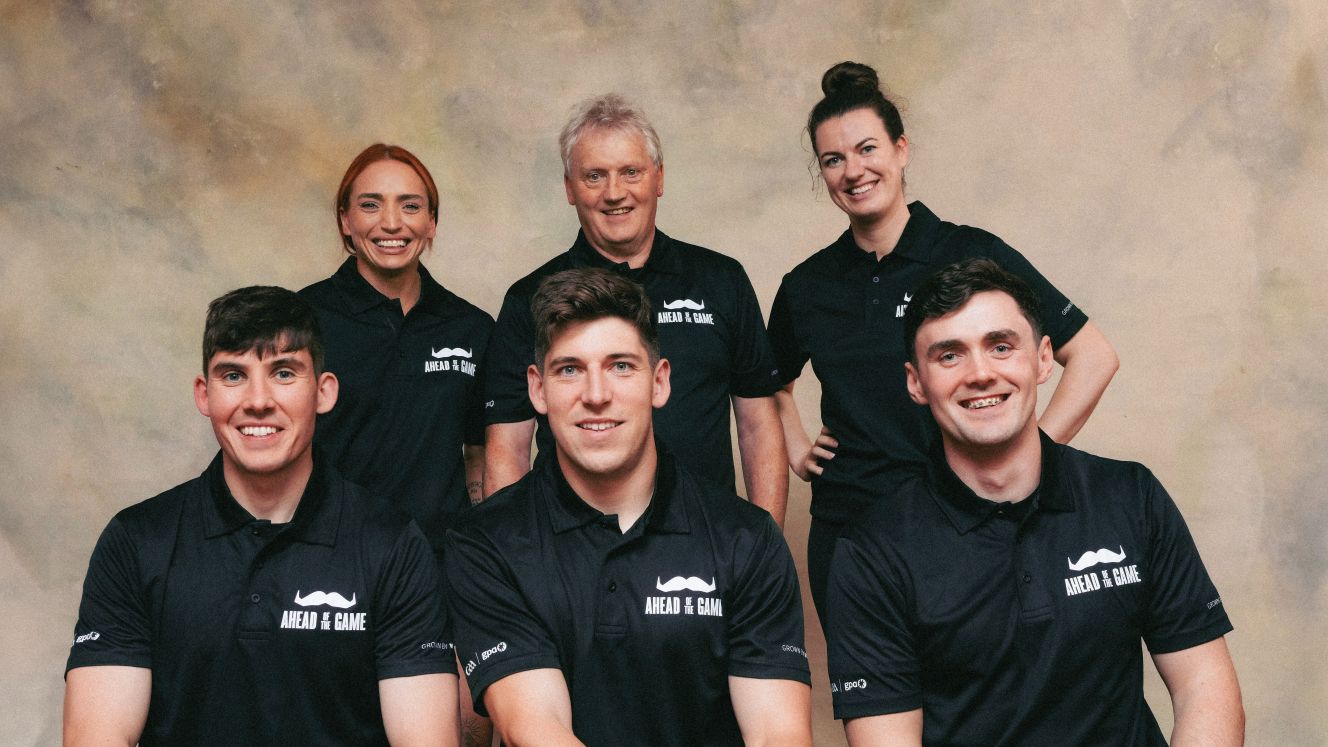 A group of three women and three men, dressed in black "Movember Ahead of the Game" shirts. Three are seated while the others stand behind them, all smiling.