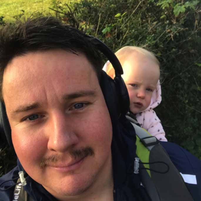 Photo of a man with a dazzling moustache, smiling to camera, carrying his infant on his back.