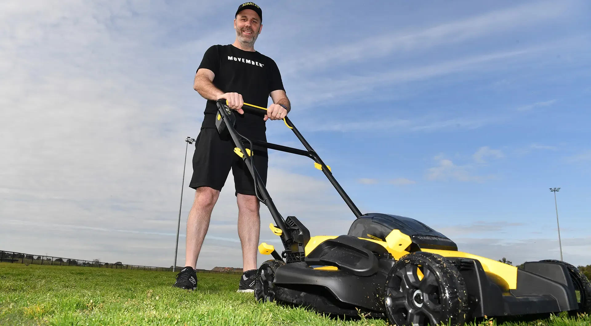 A man in Movember-branded attire, pushing a lawnmower.