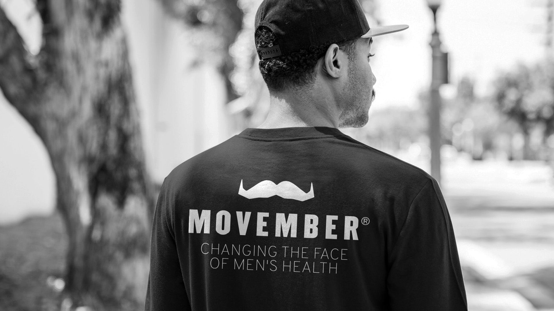 Black and white photo of a man sporting stylish Movember merchandise. The photo shows the back of the man's t-shirt, which bears large white letters that say: "Movember. Changing the face of men's health"