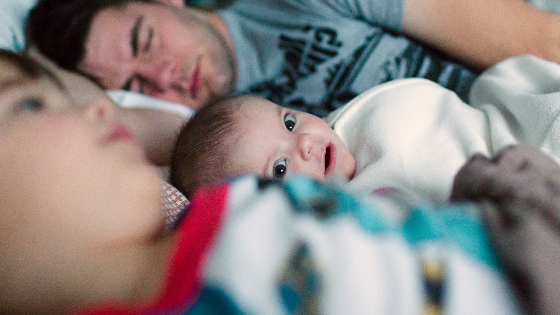 Photo of infant looking to camera betwen two sleeping parents.