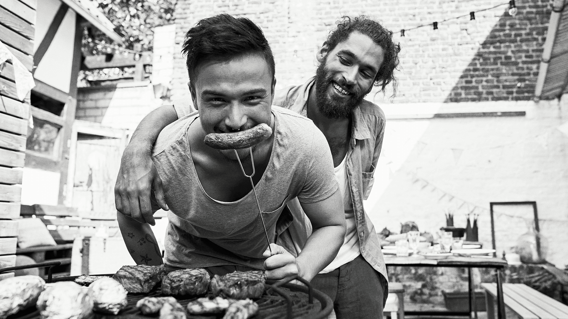 Two guys at a BBQ holding a fork with a sausage on it.