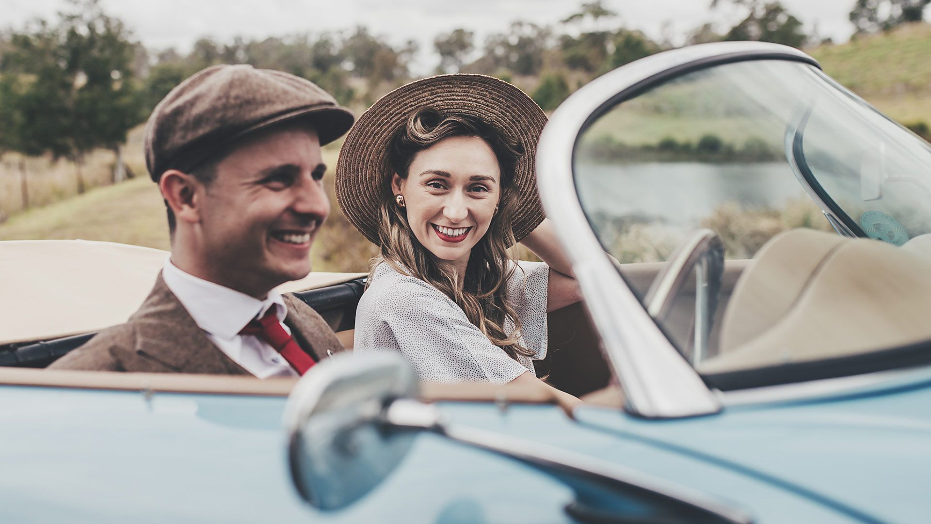 A photo of a superbly well-dressed couple, smiling from a classic car.