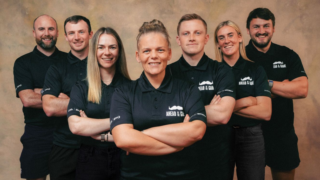 A group of three women, and four men, all wearing black "Movember Ahead of the Game" shirts. They are standing with arms crossed and smiling at the camera.