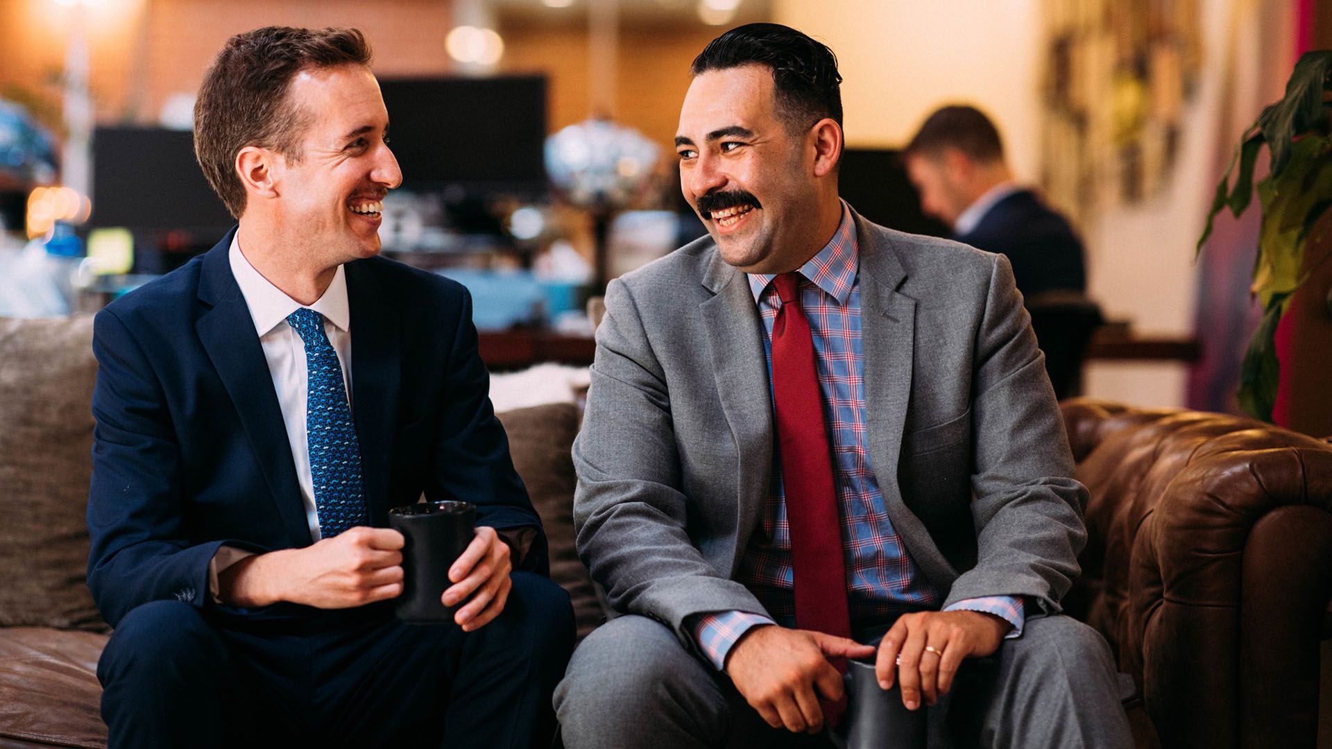 Two men in suits, bearing with moustaches, sitting and laughing in an office. 