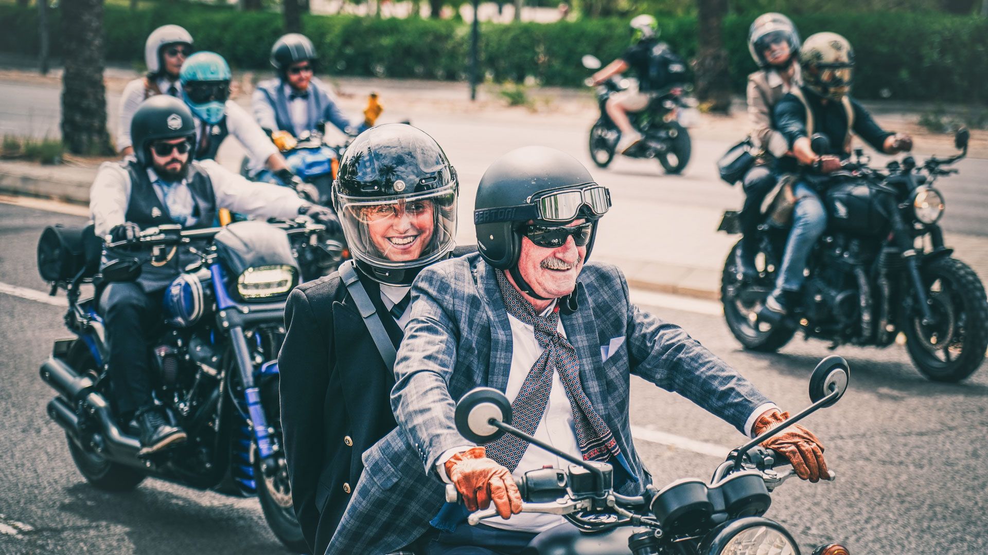 Motorcyclist ride along the road. Older man drives with younger lady on back. Other motorcyclist around them.