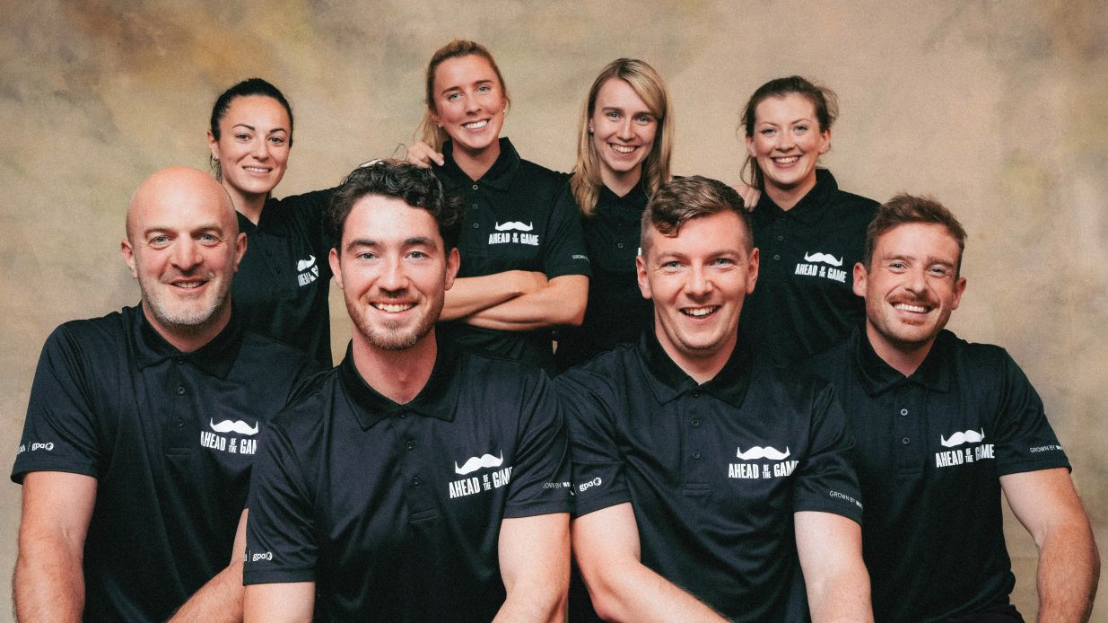 A group of four women and four men, dressed in black "Movember Ahead of the Game" tshirts. The men are seated while the women stand behind them all smiling.